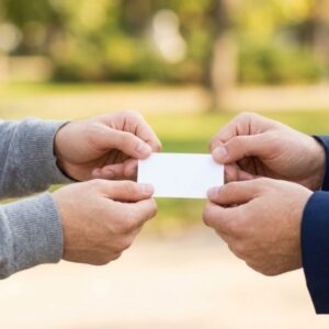 Hands exchange a blank white business card outdoors in natural daylight with a soft green blur background.